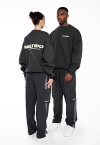 Felpe oversized nere con la scritta "MULTIPLY" stampata sulla parte anteriore e posteriore, abbinate a pantaloni cargo coordinati con tasche e dettagli grigi.