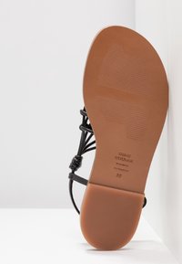 Sandale en cuir noir avec une semelle plate, présentant un dessous lisse marron, des bretelles tissées et un branding subtil sur la semelle intérieure.