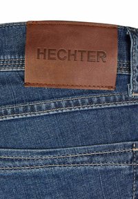 Jean en denim de couleur bleu foncé, avec une patch en cuir marron portant l'étiquette "HECHTER" à la taille. Détails de couture visibles et texture.