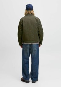 Person med langt hår iført en marineblå kasket, olivengrøn jakke, løse blå jeans og brune sko, der står foran en hvid baggrund.