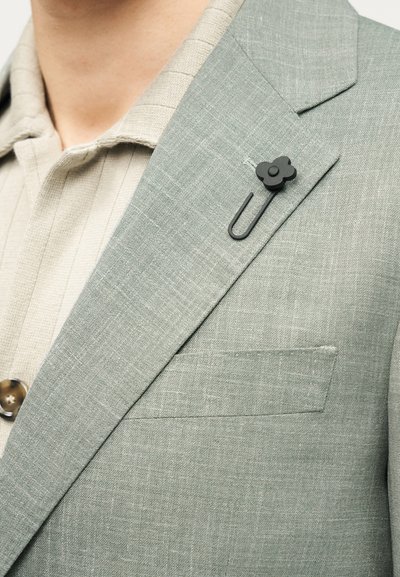 Ανοιχτό πράσινο λινό σακάκι με δομημένο πέτο, που διαθέτει μαύρο φλοράλ καρφίτσα και τσέπη με πατς, συνδυασμένο με μπεζ πουκάμισο με γιακά.
