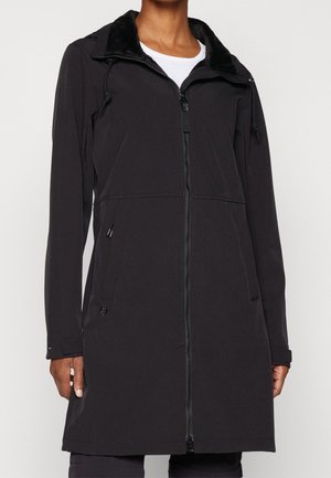 Personne portant un manteau noir mi-long zippé avec une capuche et des poches latérales, par-dessus un haut blanc, debout devant un fond uni.