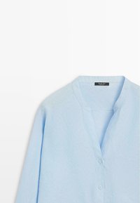 Blouse bleu clair à manches longues avec col en V et boutonnage sur le devant, en tissu texturé.