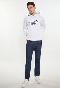 Witte hoodie met een navy "Varsity" tekst en rode sterren, gecombineerd met donkerblauwe jeans en witte sneakers. Eenvoudig, casual ontwerp.