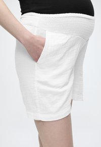 Έγκυος φοράει λευκά σορτς με ελαστική μέση και χέρι στην τσέπη, μαύρο κορμάκι, εμφανιζόμενη από τη μέση έως τη μέση του μηρού σε λευκό φόντο.