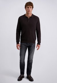 Mann trägt ein dunkles, strukturiertes langärmliges Polo, ausgebleichte schwarze Jeans und dunkle Lederloafer und steht vor einem schlichten hellen Hintergrund.