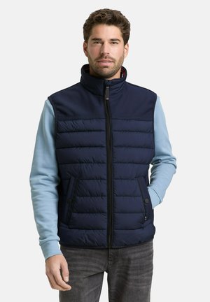 Homme portant une veste matelassée bleu marine sur une chemise à manches longues bleu clair, avec une fermeture éclair à l'avant et les mains dans les poches, debout devant un fond blanc.