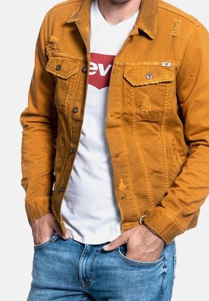 Mosterdgele spijkerjasje met gescheurde details en twee borstzakken, gecombineerd met een wit T-shirt met een rood logo, gedragen op een spijkerbroek.