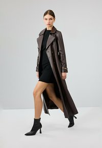 Bruine leren trenchcoat met een diepe kraag, dubbele knopen en een hoge split, gecombineerd met een strak zwart jurkje en zwarte enkellaarsjes.