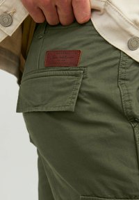 Pantaloni cargo verzi din bumbac, cu un finisaj texturat, buzunar lateral și o eticheta din piele maro cu detalii ale mărcii. Cătărarea cusăturii este vizibilă.
