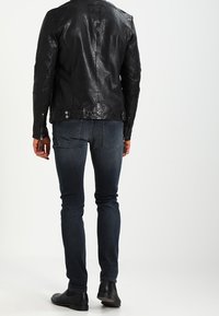Svart skinnjacka med en slät yta, med axelvaddering och sidoknappar. Bärs över mörka denimjeans och svarta skor.