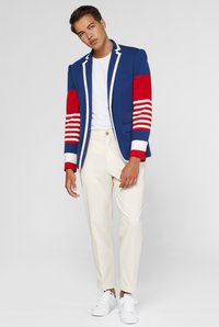 Blauwe en rode blazer met gestreepte mouwen, witte zoom, gecombineerd met crèmekleurige broeken en witte sneakers. Eenvoudig T-shirt eronder.
