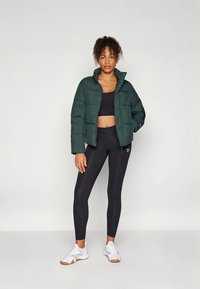 Puffer jacket in dark green, cropped design, featuring a button closure and quilted texture. Worn over a black top and paired with black leggings and white sneakers.