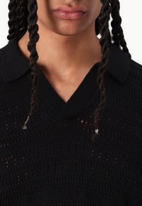 Polo shirt tricotat negru, cu un model texturat și un guler în formă de V. Twisturi vizibile de păr încadrează zona gâtului.