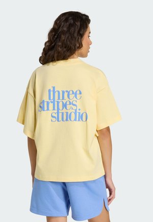 Żółta bawełniana koszulka z niebieskim napisem "three stripes studio" na plecach, zestawiona z jasnoniebieskimi szortami.