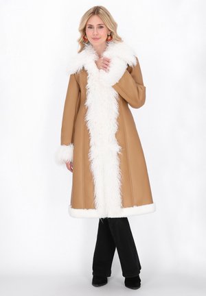 Donna che indossa un cappotto beige con bordo bianco peloso, pantaloni neri e scarpe nere, in piedi su uno sfondo bianco semplice.