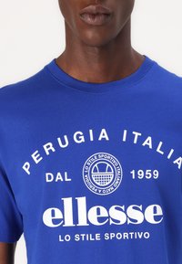 T-shirt de algodão azul com texto branco: "PERUGIA ITALIA DAL 1959" e "ellesse LO STILE SPORTIVO." Gola redonda com uma textura suave.