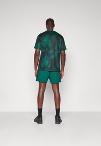 Grønnmønstret trenings t-skjorte med rund hals, paret med matchende grønne shorts. Fottøyet er svarte joggesko, brukt med svarte sokker.