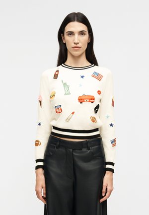 Nő fehér pulóverben, fekete csíkokkal és színes, amerikai témájú foltokkal, magas derekú fekete bőr nadrággal párosítva.