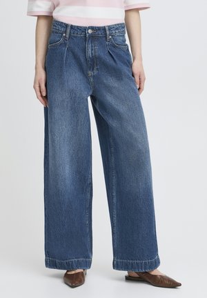 Persona che indossa jeans di denim blu a gamba larga, scarpe piatte marroni con punta appuntita e una camicia a righe rosa e bianche, in piedi contro uno sfondo semplice.