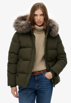 Donkergroene pufferjas met een nepbonten capuchon. Het ontwerp heeft een doorgestikt textuur en is gecombineerd met een beige gebreide trui en blauwe jeans.