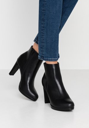 Sorte læderankelstøvler med blokhæle, båret med blå denimjeans, vist mod en ensartet lys baggrund.