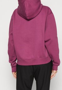 Purple hoodie with a kangaroo pocket, ribbed cuffs and hem, and a soft texture. Worn with black pants, showing the back view and hood.
