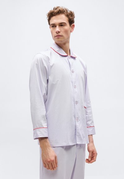 Νεαρός άνδρας που φοράει σετ πιτζάμας σε ανοιχτό μωβ χρώμα με κόκκινο περίγραμμα στον γιακά και τα μανίκια, όρθιος και κοιτάει προς το πλάι μπροστά σε λευκό φόντο.