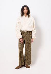 Licht ivoorkleurige oversized jas met twee zakken, gecombineerd met bruin panterprint wijde broek en bruine sandaal met meerdere banden.