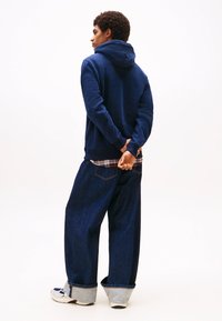 Navy blue hoodie with a hood, paired with loose-fitting dark denim jeans featuring a light blue cuff and plaid shirt underneath. White sneakers.