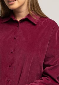 Bordowa koszula z korduroju z zapięciem na guziki z przodu, z ostro zakończonym kołnierzykiem i długimi rękawami, charakteryzująca się miękką teksturą i sześcioma pasującymi guzikami.