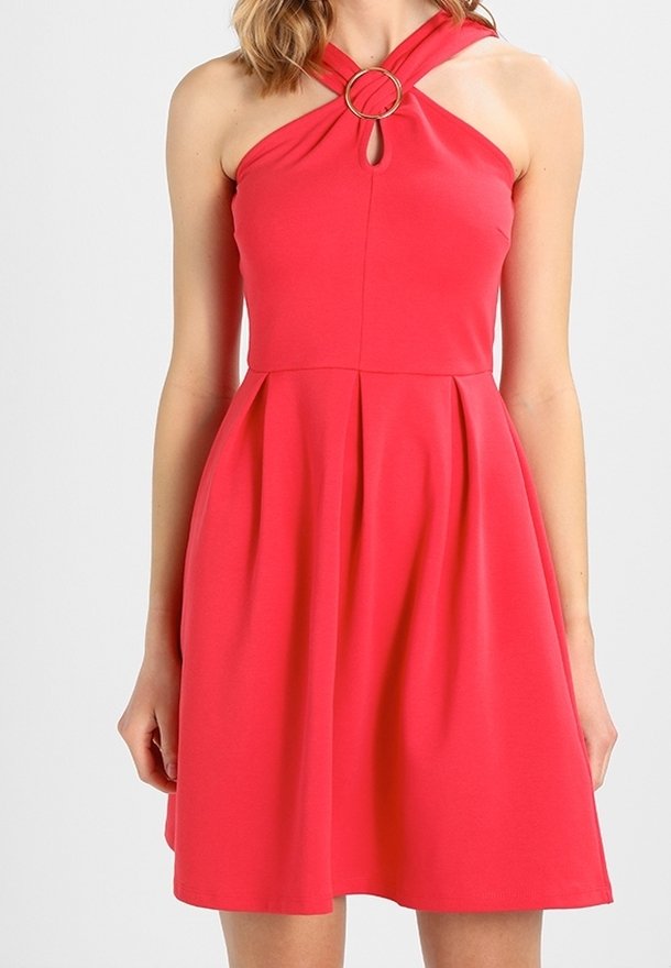 Femme portant une robe rouge sans manches avec un col halter croisé devant et un détail d’anneau métallique rond.