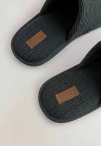 Chaussons tissés gris foncé avec une surface texturée, avec un patch en cuir marron sur chacun. Le design est simple avec des orteils ouverts.