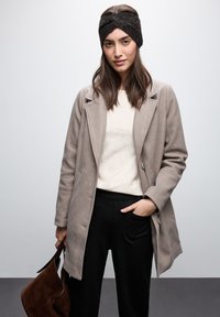 Cappotto in misto lana beige con colletto a rever e dettaglio dei bottoni, abbinato a un maglione crema e pantaloni neri. Gli accessori includono un cerchietto scuro.