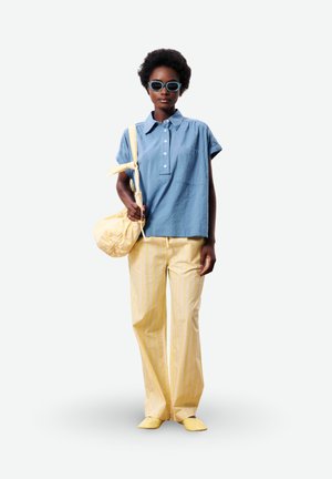 Femme portant une chemise bleue à manches courtes, pantalon jaune rayé, sac jaune et chaussures jaunes, avec des lunettes de soleil bleues, debout sur fond blanc.