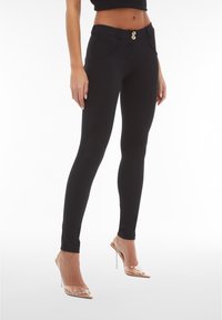 Pantaloni neri attillati con una superficie liscia, dotati di chiusura a doppio bottone e tasche laterali curve. Abbinati a scarpe con tacco trasparente.