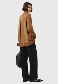 Top lungo a maniche lunghe in satin marrone con vestibilità rilassata e polsini a coste, abbinato a pantaloni neri a vita alta e gamba larga e scarpe nere flat.