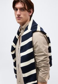 Νεαρός άνδρας με καστανά μαλλιά, που φοράει ένα μπεζ πουκάμισο με κουμπιά και ένα μπλε σκούρο και λευκό ριγέ πουλόβερ περασμένο πάνω από τους ώμους του.