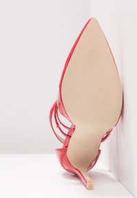 Escarpin en cuir verni rouge avec un bout pointu et un fin stiletto. Présente des brides croisées et une semelle lisse et texturée.
