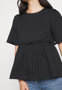 Schwarze Baumwollbluse mit kurzen, gepufften Ärmeln. Verfügt über einen gerafften Taillenbereich und eine A-Linien Silhouette. Glatte Textur, klassischer runder Ausschnitt.