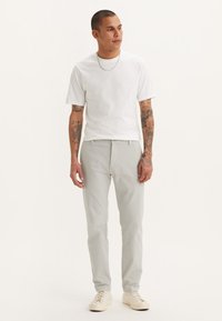 Pantalones gris claro de corte recto, combinados con una camiseta blanca de manga corta. La tela parece suave, con tatuajes visibles en los brazos. Zapatos blancos.