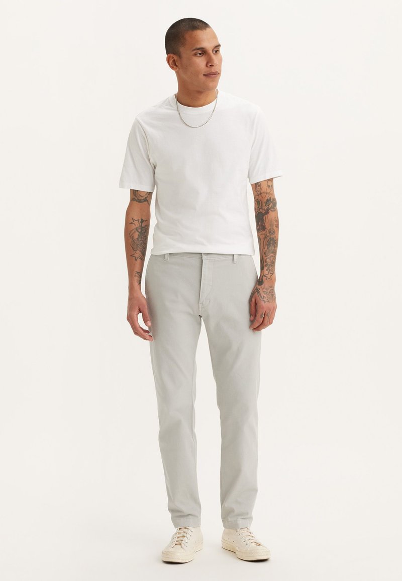 Pantalones gris claro de corte recto, combinados con una camiseta blanca de manga corta. La tela parece suave, con tatuajes visibles en los brazos. Zapatos blancos.