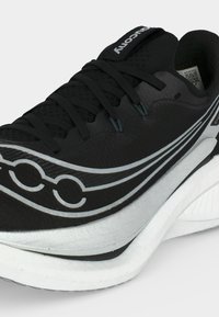 Κοντινό πλάνο ενός μαύρου αθλητικού παπουτσιού με μαύρα κορδόνια, λευκή ενδιάμεση σόλα και ασημένιο λογότυπο στο πλάι, που δείχνει υφή από διχτυωτό υλικό και επενδυμένο γιακά.