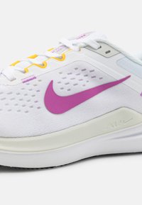 Zapatilla deportiva blanca con el swoosh morado de Nike, parte superior de malla texturizada, acentos amarillos en los cordones y una suela intermedia de color claro con un detalle de logo.