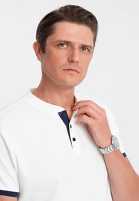 Bílá polo košile s námořnickým límcem a knoflíky. Model má krátké vlasy a nosí stříbrné náramkové hodinky, na neutrálním pozadí.