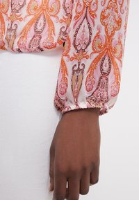 Genomskinlig blus med paisleymönster i orange och rosa nyanser. Långa, rynkade ärmar med elastiska manschetter. Vit bakgrund.