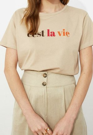Beiges T-Shirt mit „cest la vie“ in braunen, roten und orangenen Buchstaben, in hoch taillierte beige Hosen mit Doppelknöpfen gesteckt.
