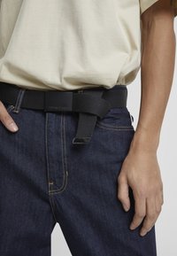 Črn mrežasti pas z pravokotno mat sponko, nosi se čez temne jeans hlače. Tkanina je trpežna, s vidnimi detajli šivanja.