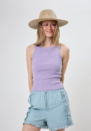 Jeune femme portant un chapeau de soleil tissé, un top lavande sans manches et un short rayé bleu clair avec des volants, souriante et regardant sur le côté.