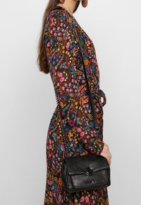 Černá kožená crossbody taška s klopou, vzorovanou texturou a detaily z kovu, nošená přes šaty s květinovým potiskem.